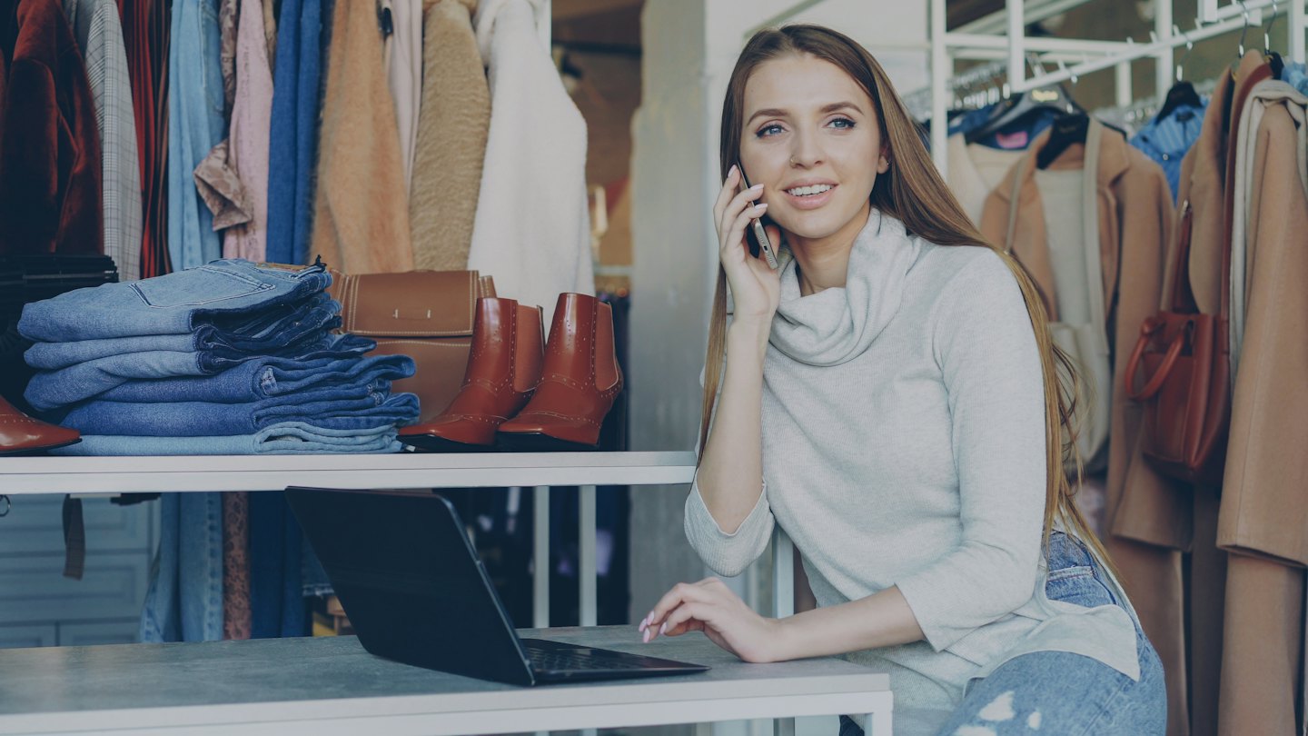 Woman managing boutique, smiling while on phone and laptop.